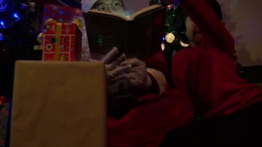 Noel Baba, Noel ağacının yanında çelenk taşıyan küçük bir çocukla kitap okuyor. Mutlu Noeller konsepti.