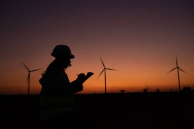 Kadın mühendisin silueti rüzgar türbini çiftliği elektrik jeneratörü istasyonunda çalışıyor ve raporu tutuyor.