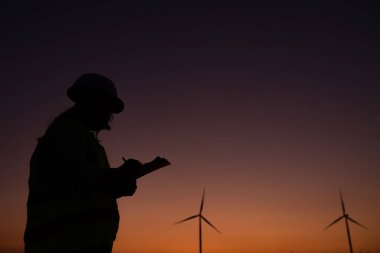 Kadın mühendisin silueti rüzgar türbini çiftliği elektrik jeneratörü istasyonunda çalışıyor ve raporu tutuyor.