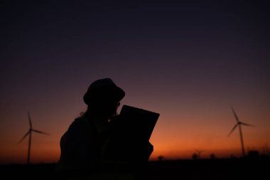 Kadın mühendisin silueti rüzgar türbini çiftliği elektrik jeneratörü istasyonunda çalışıyor ve raporu tutuyor.