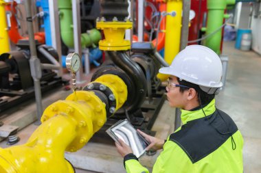 Kazan dairesinde çalışan gözlüklü Asyalı mühendis ısıtma sistemi ekipmanlarının teknik verilerini kontrol ediyor.