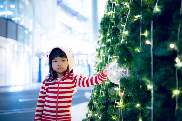 在圣诞树旁快乐地戴着圣诞帽的可爱的亚洲小女孩的画像 — 图库照片