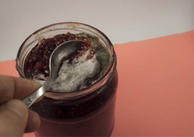Ahududu reçeli kavanozundaki beyaz ve gri küfler. Beyaz-pembe arka planda üstten çekilmiş bir fotoğraf.