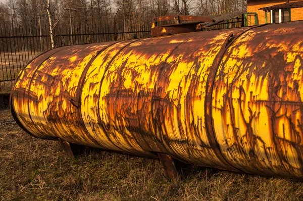Big, yellow, rusty tank on nature background and red brick house behind.