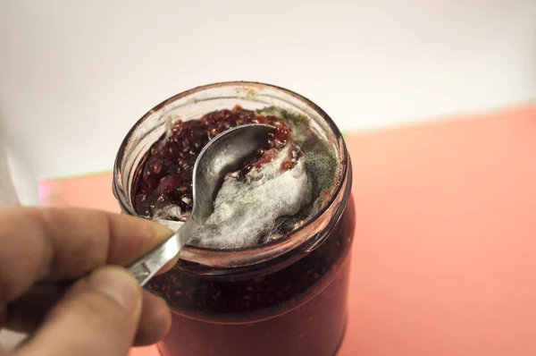 Sarı ve gri küf iplikleriyle kaplı ahududu reçeli kapağı olmayan bir kavanoz. Elinde kaşıkla reçelden küf toplayan bir el görebilirsin. Fotoğraf beyaz-pembe arka planda..