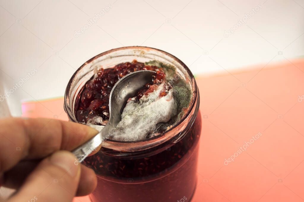 A jar without a lid with raspberry jam, which is covered with threads of yellow and gray mold. You can see a hand with a spoon raking mold from the jam.  Photo on white-pink background on the side.