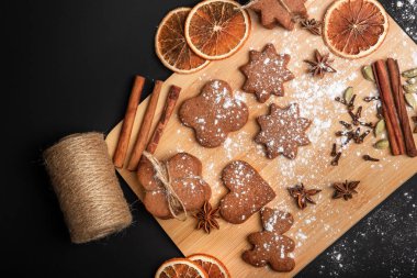 Siyah arka plana karşı ahşap bir tahtanın üzerinde baharatlı zencefilli kurabiye. Tarçın çubukları, portakal dilimleri, yıldız anason, karanfil, kakule ve güzel bir kompozisyonda pudra şekeri. Tatil atmosferi. Zencefilli kurabiyeler.