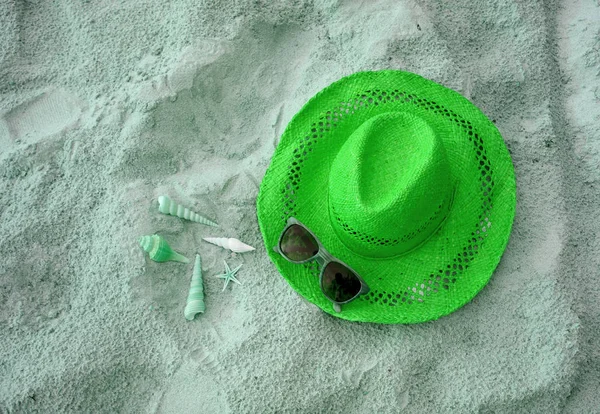 Straw hat with many types of little seashells and sunglasses on the sandy beach in green color tone