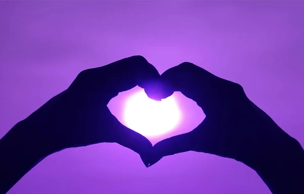 Silhouette of woman's hand posing LOVE HEART sign against the shiny sun on vibrant purple sky