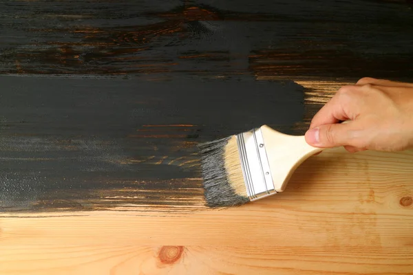 Hand holding paintbrush painting the surface of wooden furniture with dark grey paint