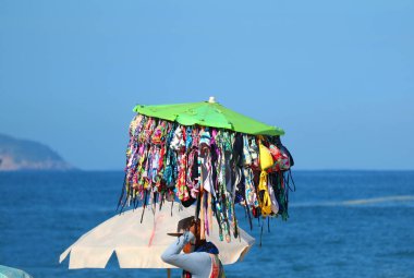 Mayo satıcısı elinde bir sürü bikiniyle sahilde yürüyor. Arka planda okyanus var.
