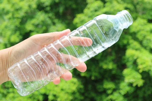 Adamın eli, bulanık yeşil yapraklara karşı boş bir plastik şişe içme suyu tutuyor.