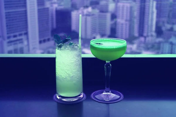 Two different type of cocktails in vibrant green color on the rooftop bar\'s table with blurry skyscrapers view in background