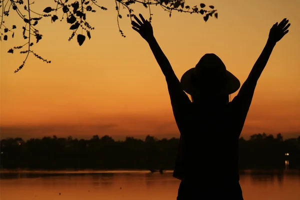 一位年轻女子在湖岸迎着美丽的橙色日落天空举起双臂的轮廓 — 图库照片