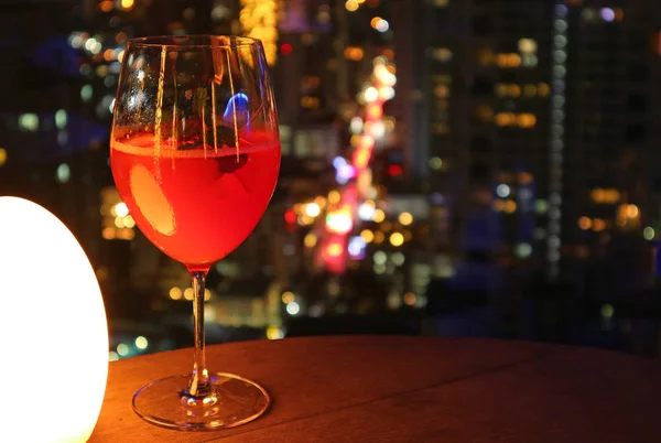 Glass of vibrant red cocktail on the rooftop bar\'s table with urban night view bokeh in background