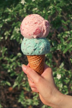 Vertical image of melting two scoops Ice cream cone in hand with blurry green trees in background clipart