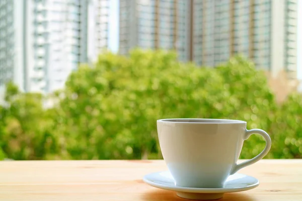 A cup of hot drinks on the window side wooden table with blurry green foliage and high buildings in background