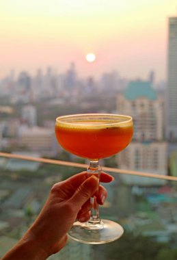 Arka planda ufuk çizgisi üzerinde gün batımının bulanık olduğu bir bardak kokteyli tutan bir kadın eli.