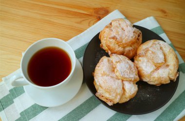 Bir tabak Fransız Kremalı Puf ya da Choux a la Creme ve bir fincan sıcak çay ahşap masada servis edilir.