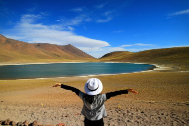 Female tourist raising her arms admiring the amazing deep blue lagoon Laguna Miniques, located in the altiplano of Antofagasta region, Chile clipart