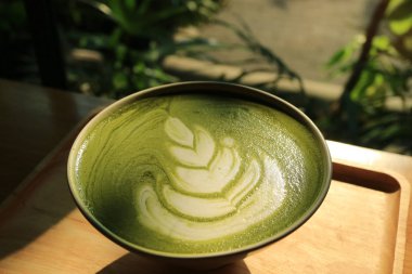 Bir fincan sıcak Japon kibrit yeşil çay arkaplanda bulanık bitkilerle gün ışığında.