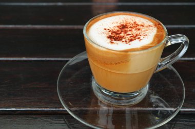 Koyu renkli ahşap masada servis edilen şeffaf fincanda sıcak kapuçino kahvesi.