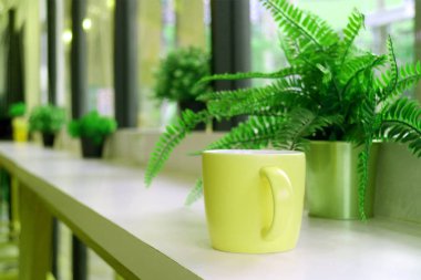 Sarı kahve kupası, eğreltiotu saksısı ve pencere kenarında uzun bir masa.