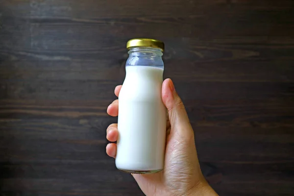 Hand holding glass bottle of milk against dark brown wooden wall