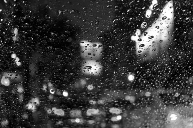 Geceleri arabanın ön camındaki yağmur damlalarından görülen bulanık sokak lambaları ve kuyruk lambalarının tek renkli fotoğrafı