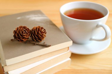 Bir fincan sıcak çayın bulanık arka planına sahip bir yığın kitabın üzerindeki gül kozalakları.