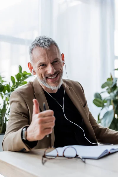 selective focus of happy businessman in earphones showing thumb up in office