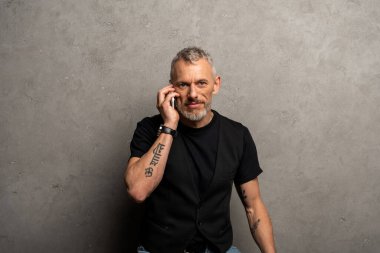 Sakallı ve dövmeli bir adam gri üzerine akıllı telefondan konuşuyor. 