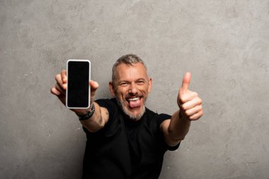 Seçici odak noktası dilini dışarı çıkaran ve gri ekranda parmağını gösteren akıllı telefonu tutan mutlu bir adam. 