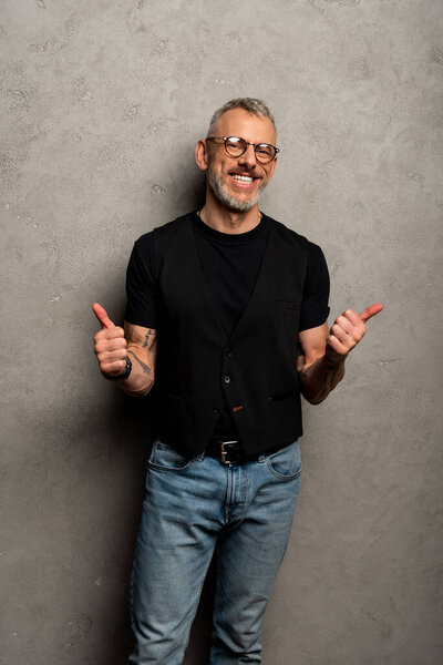 cheerful man in glasses showing thumbs up on grey 