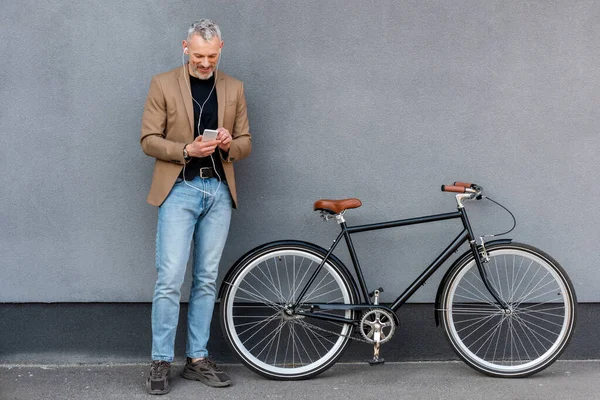 happy businessman in earphones listening music while holding smartphone near bicycle outside