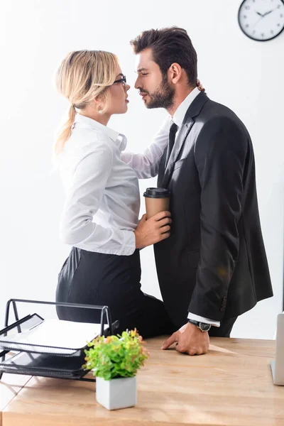 Вид сбоку на сексуальную бизнесвумен, держащую кофе и обнимающую бизнесмена в офисе — стоковое фото
