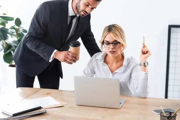 Empresário segurando café enquanto colega trabalhando com laptop — Fotografia de Stock