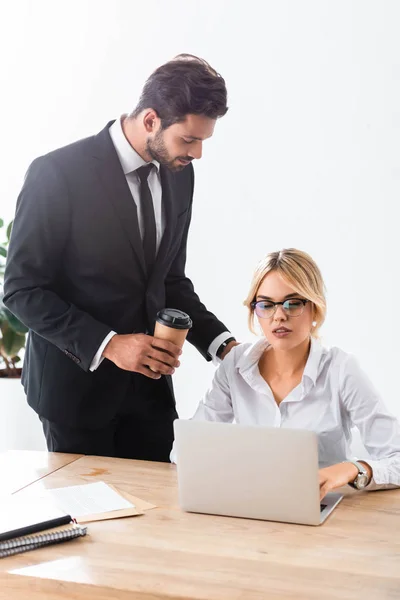 Empresario con café mirando a colega trabajando con portátil - foto de stock