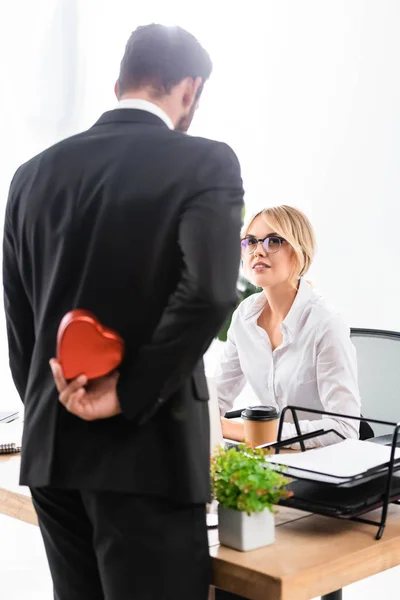 Enfoque selectivo de la secretaria sonriente mirando a hombre de negocios ocultando regalo - foto de stock