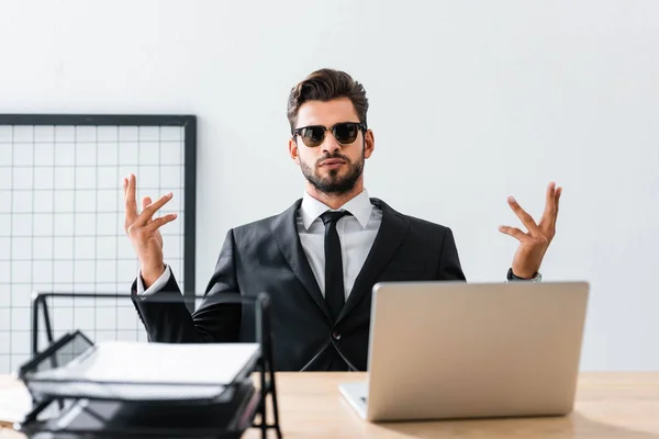 Bonito homem de negócios em óculos de sol gesticulando na mesa de escritório — Fotografia de Stock