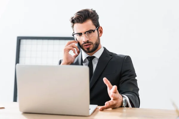 Empresario hablando en teléfono inteligente y mirando a la computadora portátil en la oficina - foto de stock