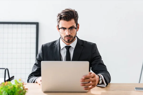 Bonito homem de negócios olhando para a câmera enquanto usa laptop — Fotografia de Stock