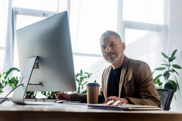 Selektive Fokussierung des bärtigen Geschäftsmannes auf Kamera in der Nähe von Coffee to go und Computermonitor im Büro — Stockfoto