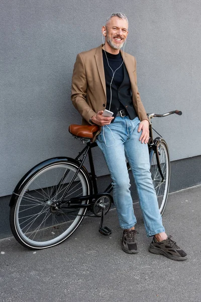 Alegre hombre de negocios en auriculares escuchando música mientras sostiene el teléfono inteligente y de pie cerca de la bicicleta fuera - foto de stock