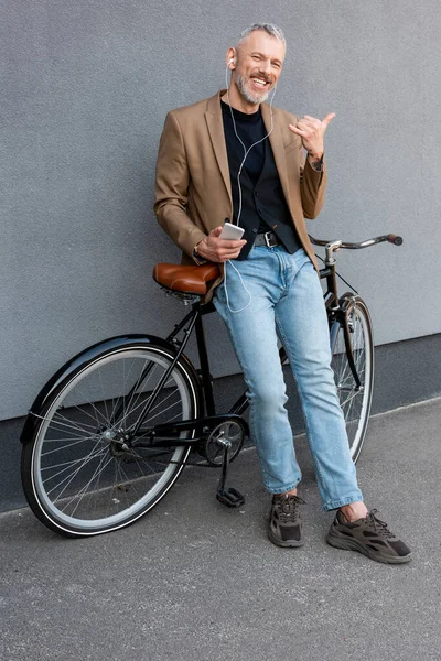 Alegre hombre de negocios en auriculares escuchando música mientras me muestra llámame gesto y sosteniendo teléfono inteligente cerca de la bicicleta fuera - foto de stock