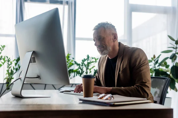 Вибірковий фокус бородатого бізнесмена, який працює біля кави, щоб піти і комп'ютерний монітор в офісі — стокове фото