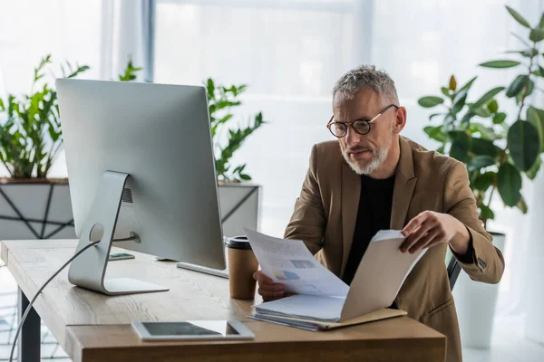 Foco seletivo do empresário barbudo em óculos olhando para documento perto de café para ir e monitor de computador no escritório — Fotografia de Stock