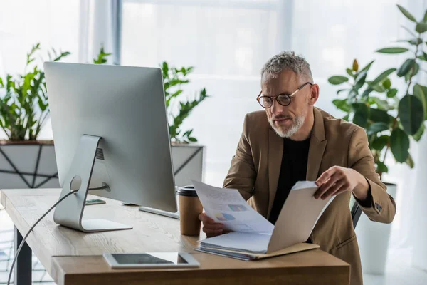 Foyer sélectif de barbu homme d'affaires dans les lunettes en regardant le document près du café pour aller et moniteur d'ordinateur dans le bureau — Photo de stock