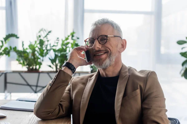 Fröhlicher Geschäftsmann mit Brille, der im Büro auf dem Smartphone spricht — Stockfoto