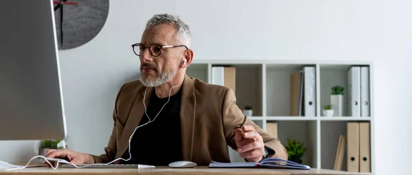 Plan panoramique de bel homme d'affaires dans les écouteurs regardant le moniteur d'ordinateur dans le bureau — Photo de stock